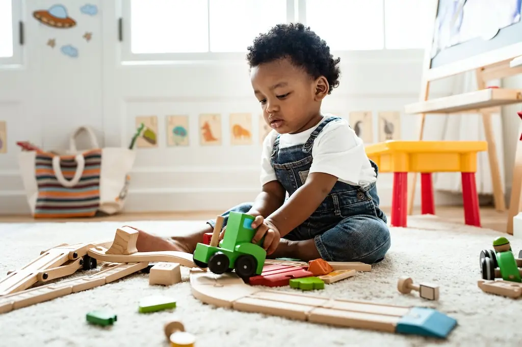 Toddler in denim dungarees pushing a green wooden train along a wooden track on a cream rug