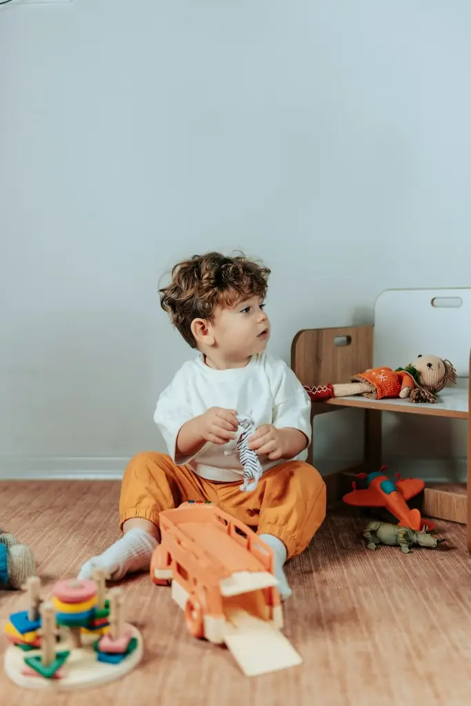 Boy holding a toy zebra beside a wooden ramp truck, a red plane and knitted dolls on a low bench behind him