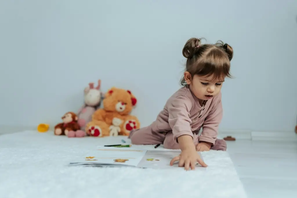 Toddler in a soft pink romper crawling towards a sticker book on a white rug, teddies propped against the wall behind her