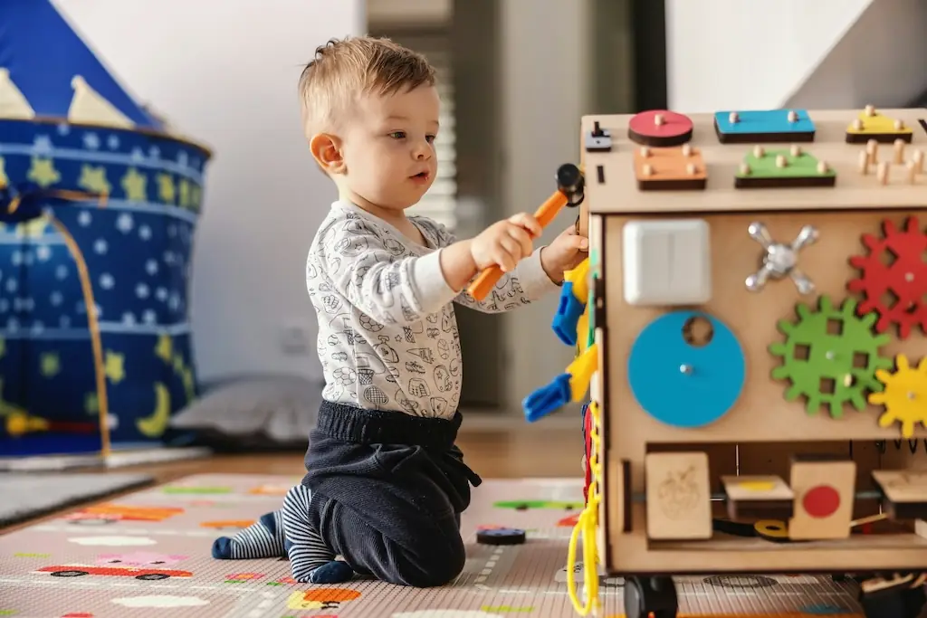 Young boy kneeling on a play mat, tapping a toy hammer against a wooden activity cube with gears and shapes