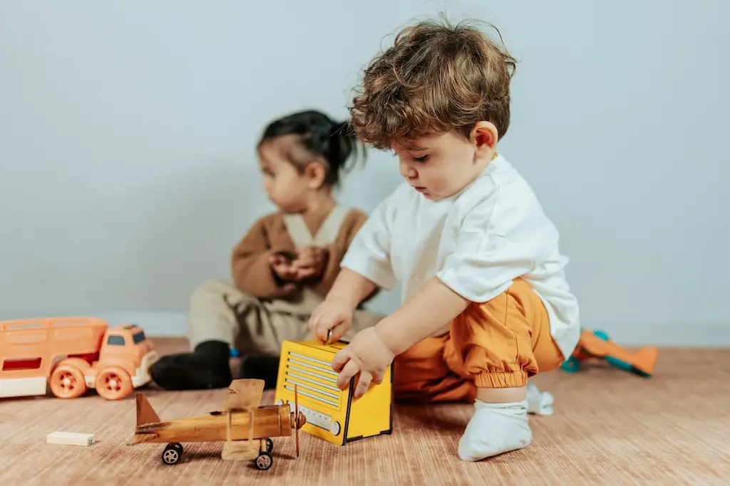 Two toddlers on a rug with a wooden aeroplane, a yellow toy radio and an orange truck between them