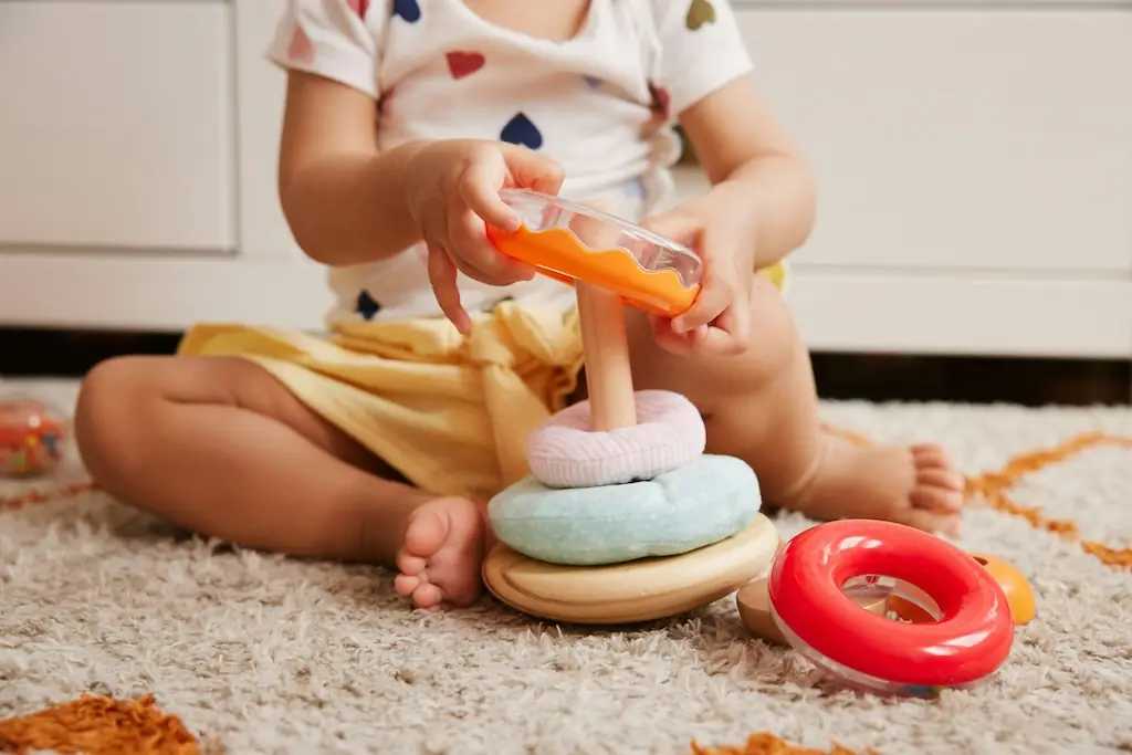 Baby sitting on a soft rug, lifting the top piece off a pastel wooden stacking-ring toy