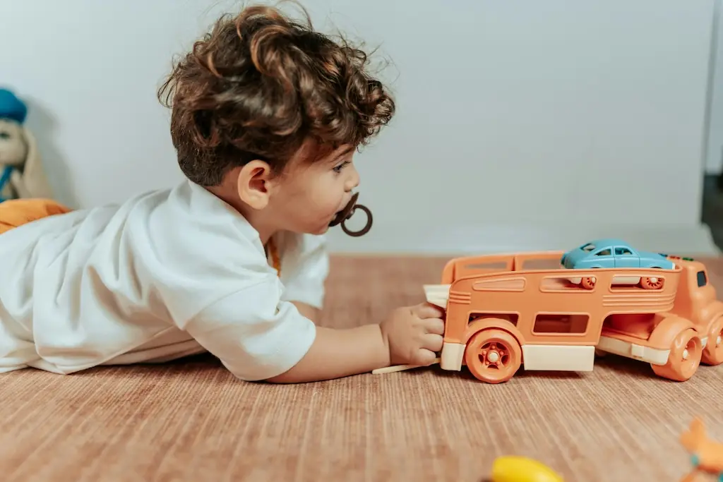 Toddler lying on their tummy reaching for an orange toy car transporter loaded with a blue car