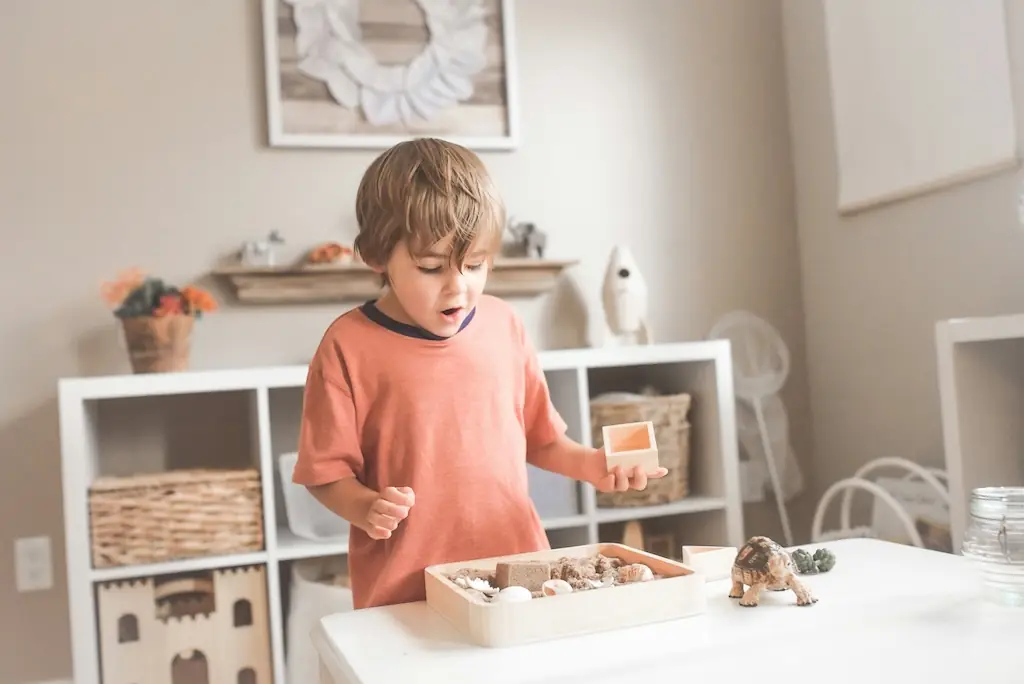 Boy in an orange t-shirt looking delighted at a wooden sensory tray with stones and a toy tortoise