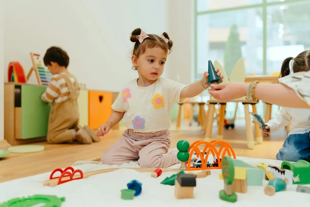 Girl in a nursery play session being handed a green wooden tree by another child, wooden train set spread out in front of them