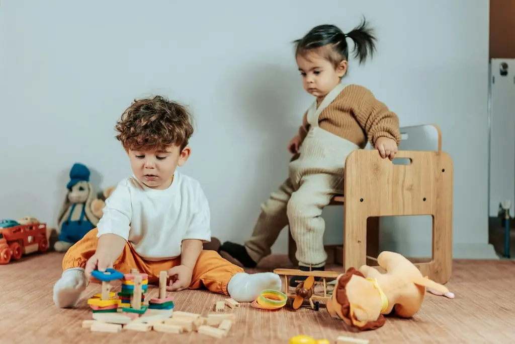 Boy sorting colourful wooden shapes on the floor while a younger sibling climbs a wooden chair behind him
