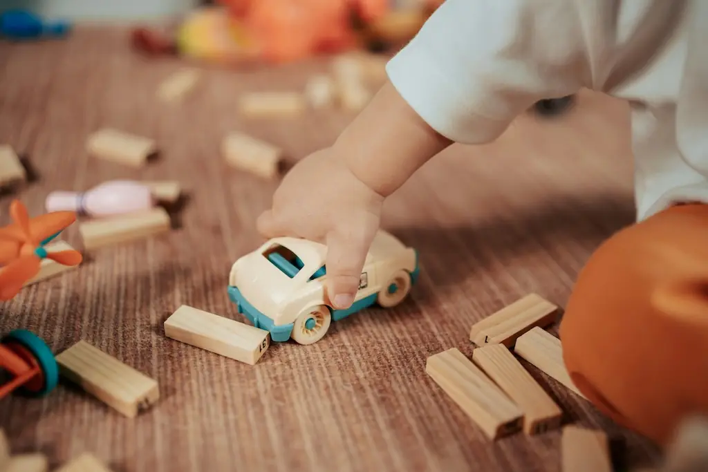 Close-up of a small hand rolling a wooden toy car between scattered wooden building blocks on a rug