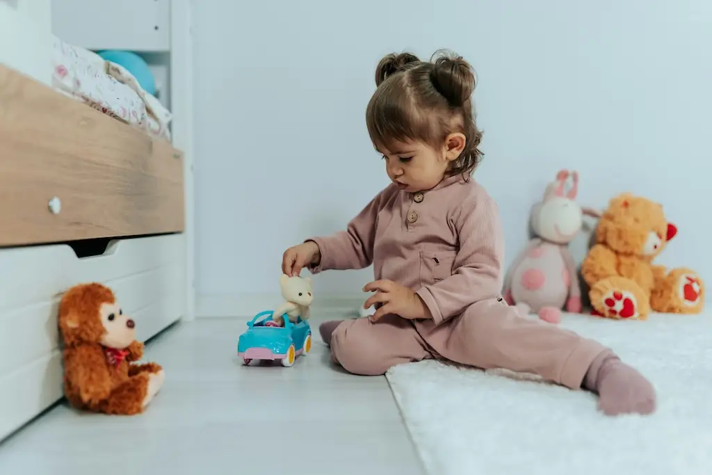 Toddler sitting beside her bed placing a small bear into a blue toy car, soft toys lined up along the wall