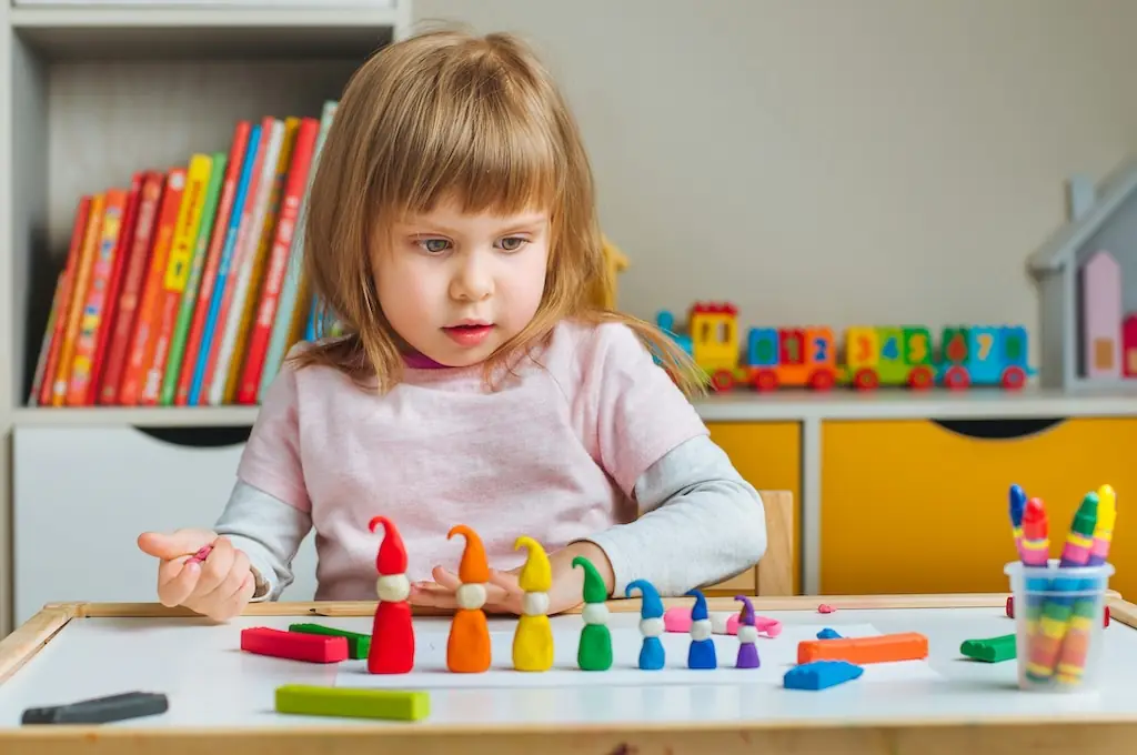 Preschool girl at a white table arranging a row of colourful play-dough gnomes she has made