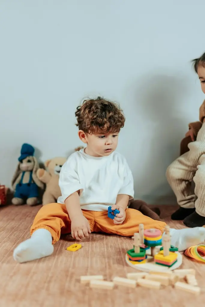 Curly-haired boy in orange trousers sitting cross-legged with a wooden shape sorter and scattered building blocks