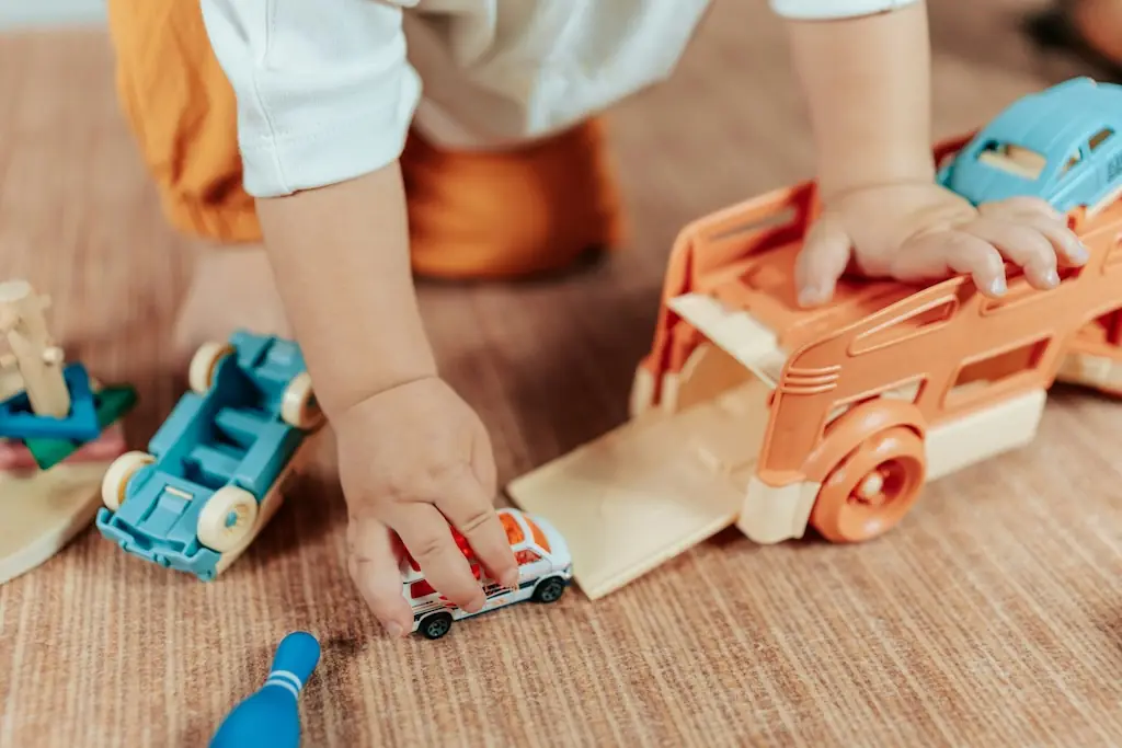Child crouched over a toy car transporter, rolling a small car down its ramp beside a blue wooden truck
